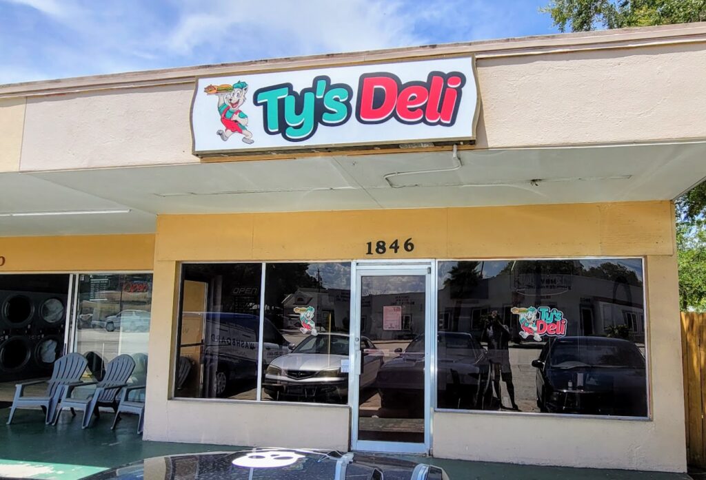 Ty's Deli front entrance off 20th street in Vero Beach Florida, cream colored walls, with lots of windows, and a sign that has a mascot with green, red, black and white colors