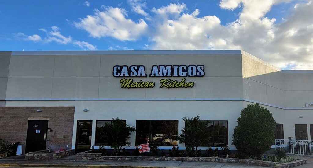 Casa Amigos Mexican Restaurant in Vero Beach mall. Cream-colored building with white trim, white and yellow sign. Entrance is landscaped with trees.