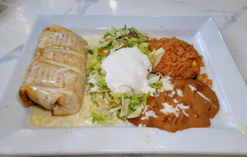 Shredded beef-filled chimichanga topped with cheese sauce, garnished with lettuce, served with sour cream, beans, and rice.