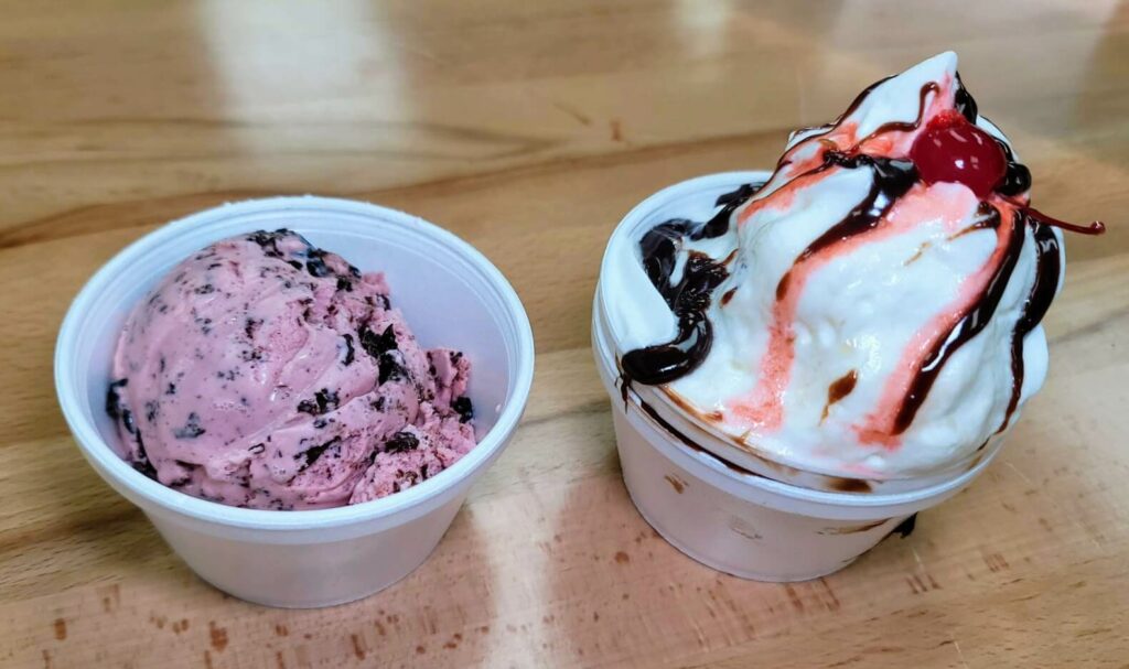 A scoop of Cherry Oblivion ice cream in a cup, and a hot fudge sundae with mint chocolate chip ice cream, topped with whipped cream and a cherry.