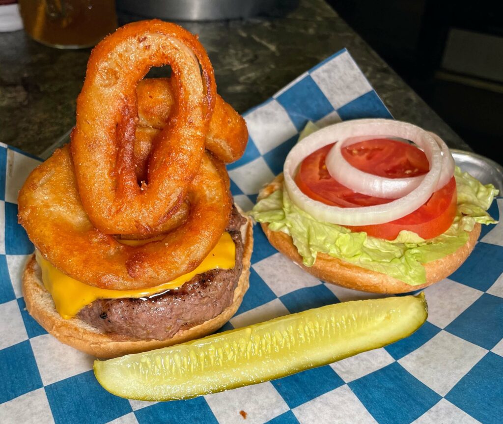 Sandy's Burger at Sandy's Grille in Sebastian, Florida, a juicy and delicious burger served with crispy onion rings, fresh lettuce and tomato, and a side of pickle spear for added flavor.