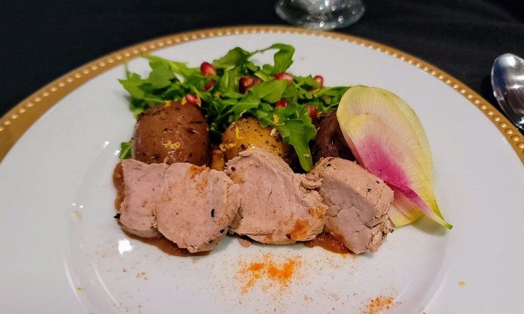 a plate of food Pork Tenderloin served with Burnt Miso Butterscotch, Confit Potatoes, Arugula Salad, and Sriracha Powder. on the indian river state college mueller campus in vero beach florida