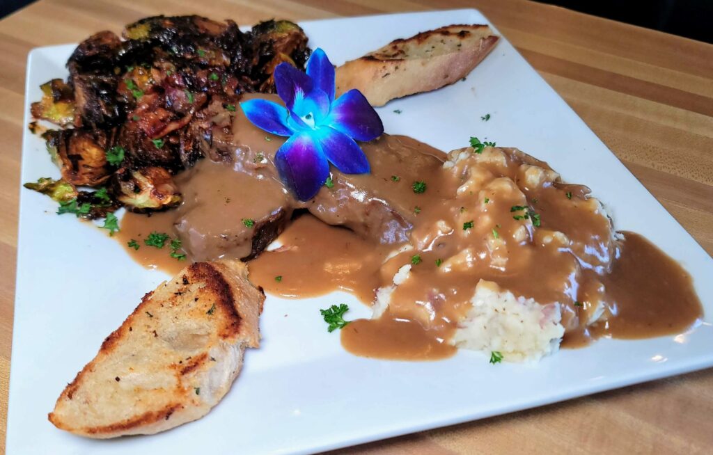 A large platter with 2 slices of Meatloaf & Mashed Potatoes topped with brown gravy, served with a side of sweet & savory charred brussels from Sacred Grounds Corner Cafe in Fort Pierce Florida