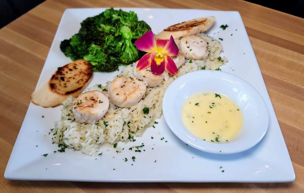 Jumbo seared sea scallops served over a bed of rice topped with a pink orchid flower, served with broccoli, crustini, and a side of beurre blanc sauce from Sacred Grounds Corner Cafe in Fort Pierce Florida