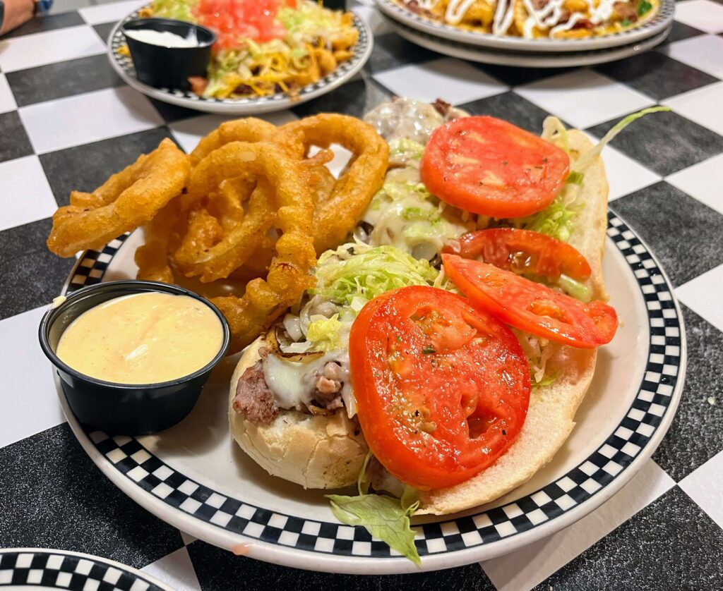 Steak hoagie Cruiser served with a side of onion rings, the sandwich has steak and is also topped with onions, cheese, lettuce and tomato at Mrs Mac's Fillin Station restaurant located in Vero Beach Florida