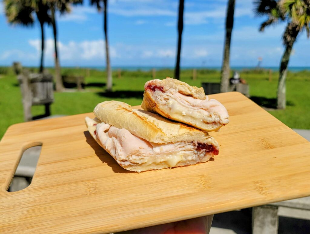 a turkey, cranberry, and brie panini sandwich as prepared by Polo Deli located in vero beach florida displayed on a cutting board at Humiston Park with an ocean view in the background