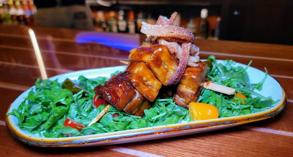 BBLT features Bourbon Glazed Cheshire Pork Belly Kebabs, paired with Orange Blossom & Vanilla Bean Honey Vinaigrette dressed Arugula and a medley of Baby Heirloom Tomatoes. as prepared by Stringers Tavern & Oyster Bar in stuart florida