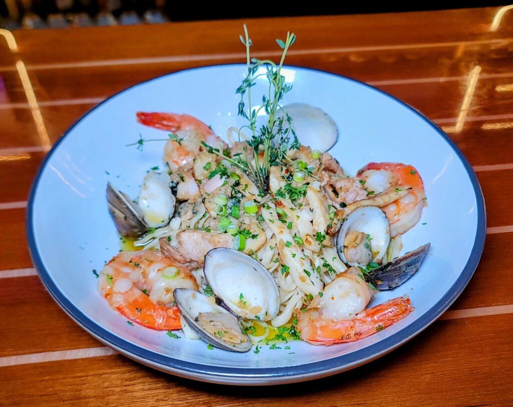 Bow to Stern Seafood Pasta, a medley of ocean-fresh ingredients, including clams, jumbo shrimp, fresh fish, and calamari, all harmoniously tossed in a flavorful blend of olive oil, fresh garlic, tomato, and green onions as prepared by Stringers Tavern & Oyster Bar in stuart florida